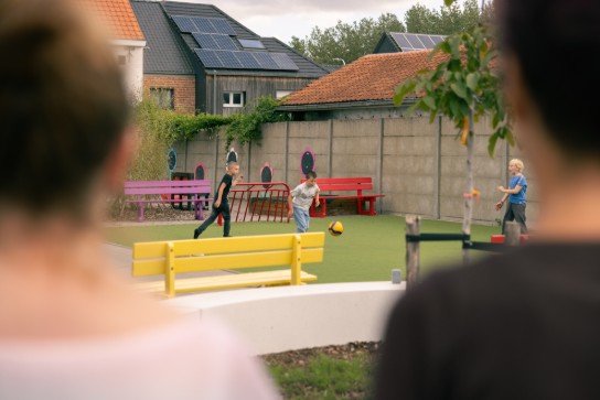 Kinderen spelen voetbal op een groen veld met kleurrijke bankjes, gezien tussen twee volwassenen in. (vergroot foto)