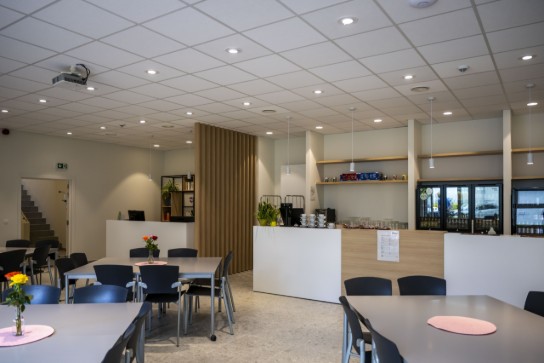 Moderne, lichte kantine met tafels, stoelen, bloemen en een bar met koffieapparatuur en koelkasten. (vergroot foto)