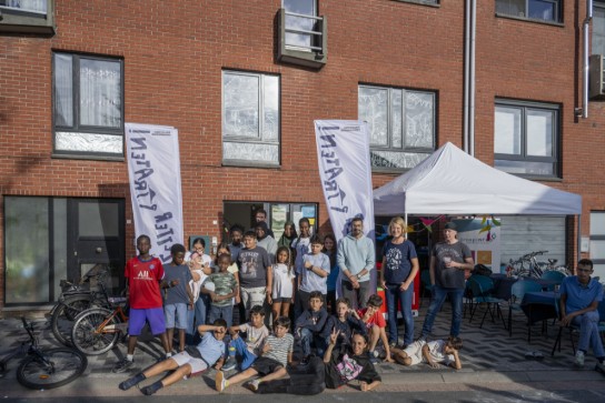Groep diverse kinderen en volwassenen poseren samen voor een gebouw met vlaggen en een witte tent op straat. (vergroot foto)